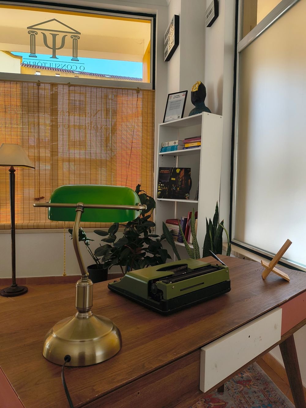 Estante com livros e plantas junto à janela, com a secretária em primeiro plano
